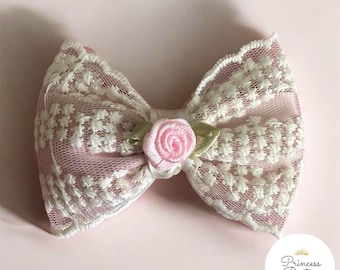 Puede incluir: Un lazo de encaje rosa y blanco con una pequeña rosa rosa en el centro. El lazo está hecho de una delicada tela de encaje y tiene una forma clásica. Un accesorio para el cabello.