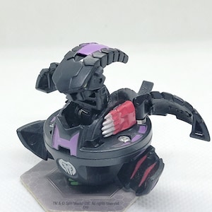 Peut inclure: Jouet Bakugan noir et violet avec des détails rouges et blancs. Le jouet est en position fermée, avec ses bras et ses jambes repliés. Le jouet est sur une carte hexagonale grise et blanche avec le texte "TM & © Spin Master Ltd. All rights reserved. E49".