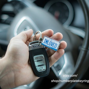 May include: A silver car key fob with a black key chain attached to a blue and silver license plate keychain that says "IMLOVED".