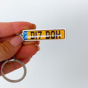 Op de afbeelding: Een zilveren sleutelhanger met een gele en blauwe kentekenplaat-hanger met de tekst "DI7 DOH".