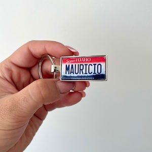 Könnte beinhalten: Ein silberner Schlüsselanhänger mit einem rot-weiß-blauen Design einer Idaho-Kennzeichen. Das Kennzeichen lautet "Scenic Idaho" und "Mauricio's Famous Potatoes".