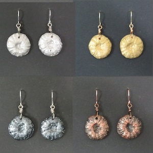 May include: Four pairs of earrings with different finishes. The earrings are silver, gold, black, and copper. Each pair features a round, textured disc with a hole in the center.