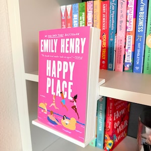 May include: A white L-shaped acrylic book display stand holding a copy of the novel Happy Place by Emily Henry. The book cover is bright pink with white text and an illustration of people swimming in a pool with yellow inflatable rings.