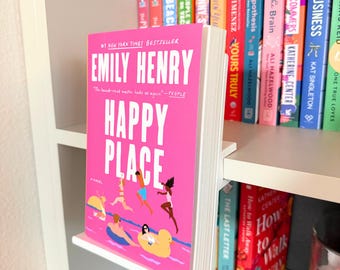 Puede incluir: Un soporte para libros en forma de L de acrílico blanco que sostiene la novela Happy Place de Emily Henry. La portada del libro es de color rosa brillante con texto blanco y una ilustración de personas nadando en una piscina con flotadores.