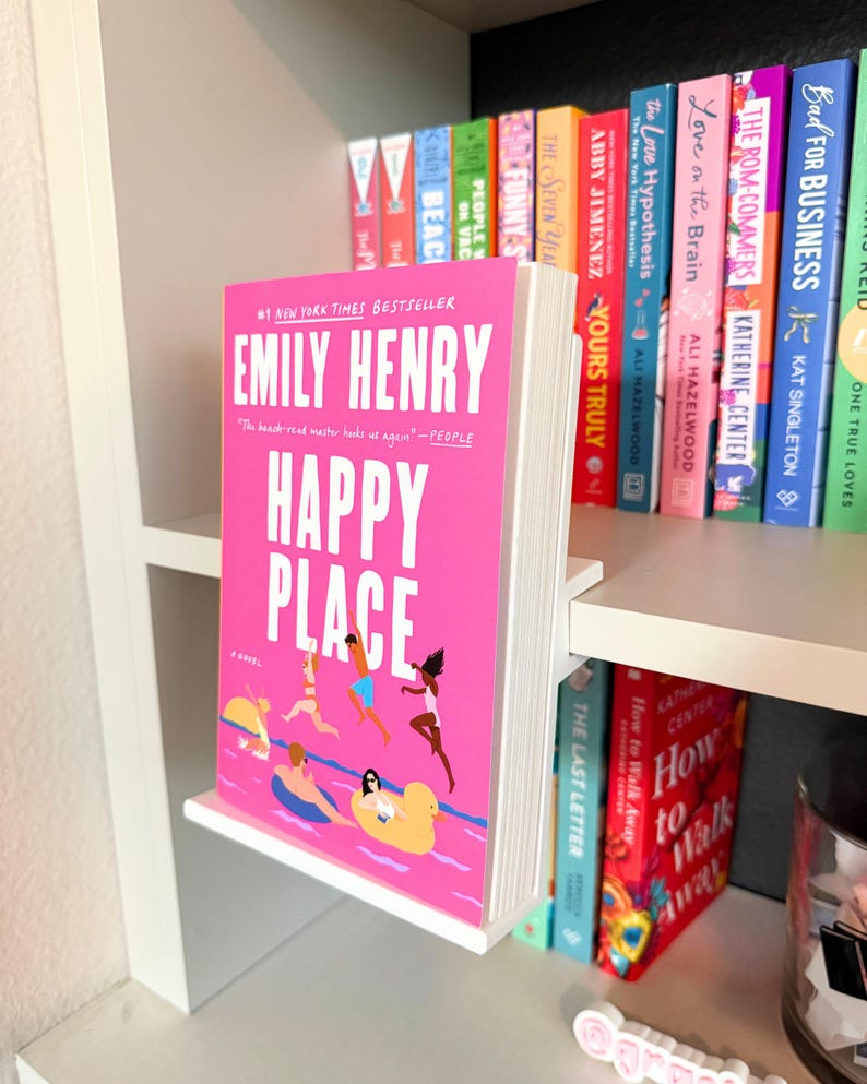 May include: A white L-shaped acrylic book display stand holding a copy of the novel Happy Place by Emily Henry. The book cover is bright pink with white text and an illustration of people swimming in a pool with yellow inflatable rings.