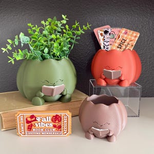 May include: Three pumpkin-shaped planters in olive green, orange, and mauve, each featuring a character holding a book. The orange planter holds patterned cards with the text "SPICE society." A rectangular orange ticket sign reads "Fall vibes BOOK CLUB."