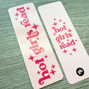 May include: Two rectangular bookmarks with rounded corners, featuring a light pink background and hot pink text. The text reads "hot girls read" in a stylized font, accented by small four-pointed star graphics. One bookmark has a black circular logo.