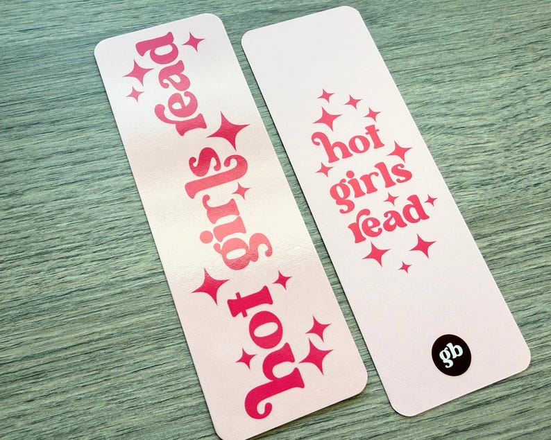 May include: Two rectangular bookmarks with rounded corners, featuring a light pink background and hot pink text. The text reads "hot girls read" in a stylized font, accented by small four-pointed star graphics. One bookmark has a black circular logo.