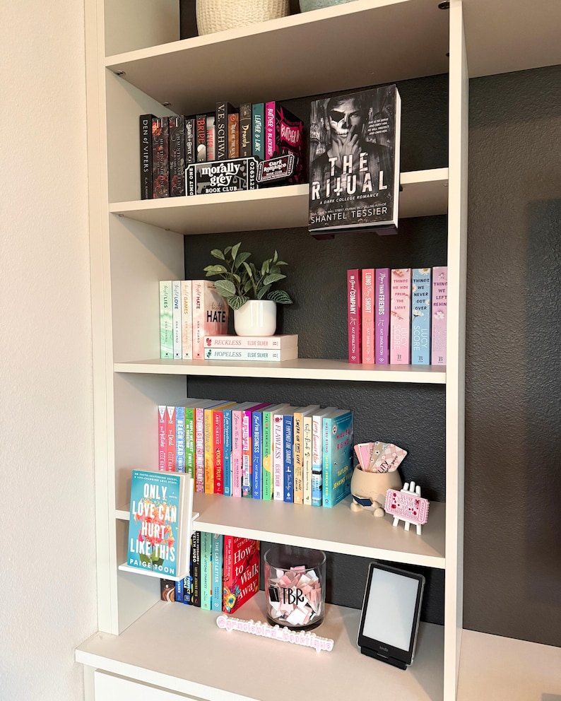 May include: A white bookshelf with five shelves containing various books, a small potted plant, a glass jar filled with paper slips labeled TBR, a digital e-reader, and decorative signs including one that reads Only Love Can Hurt Like This.