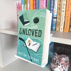 May include: A book titled Unloved by Peyton Corinne, featuring a light green cover embellished with sparkling rhinestones. The cover art displays a small open book, a dark green rectangular object, and a dark circle, all rendered in crystals.