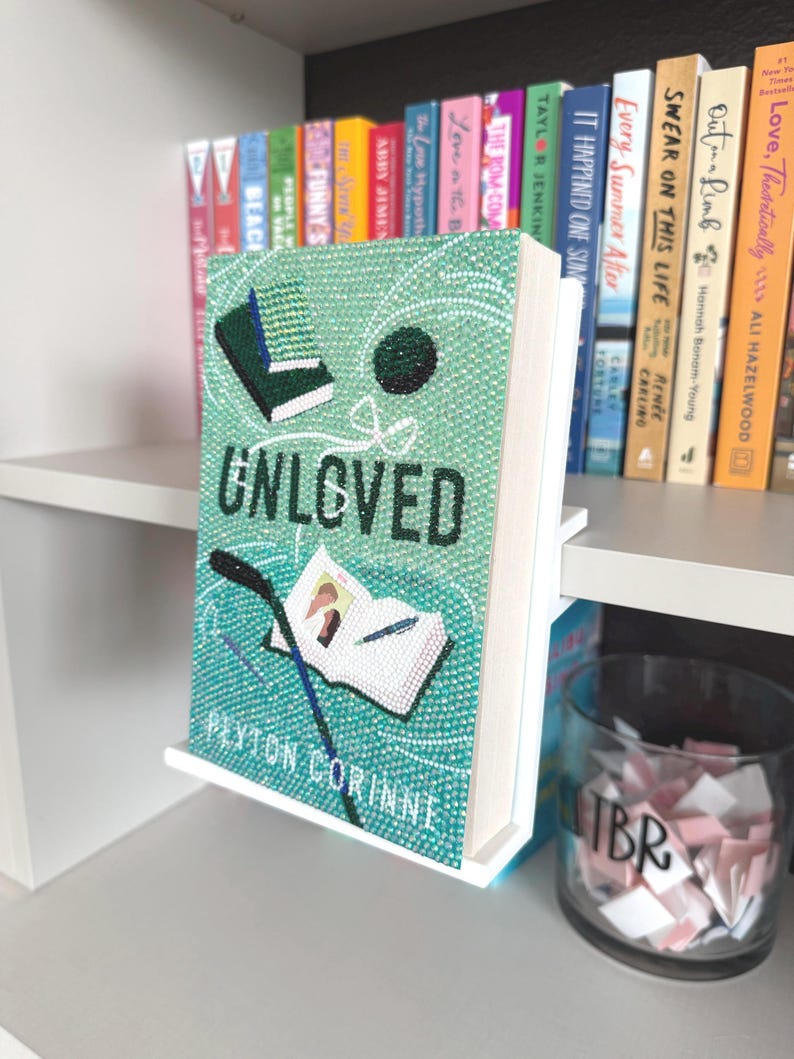 May include: A book titled Unloved by Peyton Corinne, featuring a light green cover embellished with sparkling rhinestones. The cover art displays a small open book, a dark green rectangular object, and a dark circle, all rendered in crystals.