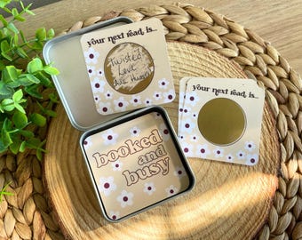 May include: A square metal tin containing cards with a cream background and white flower pattern. One card reads "booked and busy" and another "your next read is..." with a gold scratch-off circle revealing "Twisted Love".