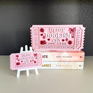 May include: A pink and white ticket-shaped sign and a smaller matching ticket on a white easel. The text reads In my bookish era BOOK CLUB LIFETIME MEMBERSHIP ADMIT ONE 02631224, decorated with red hearts, stars, and a bow graphic.