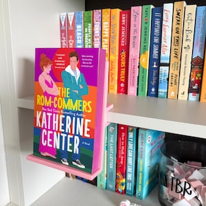 May include: A pink plastic book display stand holding the novel The Rom-Commers by Katherine Center. The book cover features a colorful illustration of two people standing against a vibrant background with bold yellow and white text.