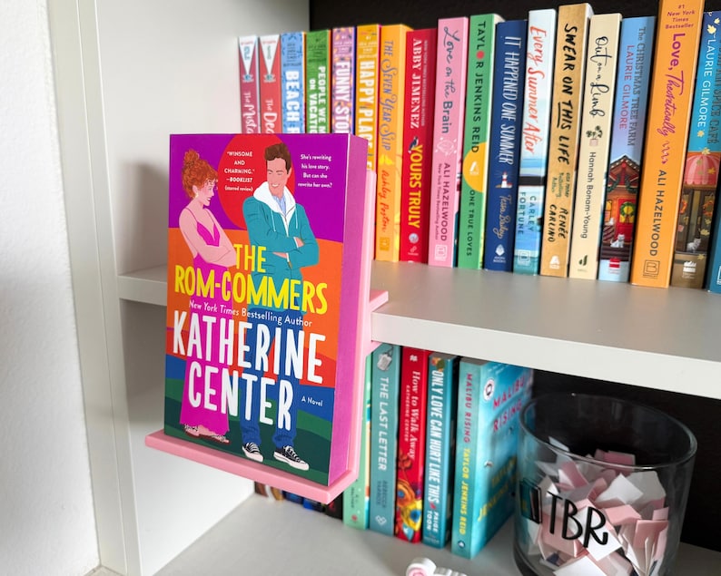 May include: A pink plastic book display stand holding the novel The Rom-Commers by Katherine Center. The book cover features a colorful illustration of two people standing against a vibrant background with bold yellow and white text.
