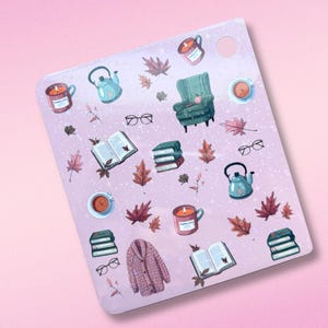 May include: A sticker sheet with a pink background featuring fall-themed illustrations. The sheet includes images of a teal armchair, books, eyeglasses, candles, teacups, kettles, and fall leaves. A pink cardigan is also featured.