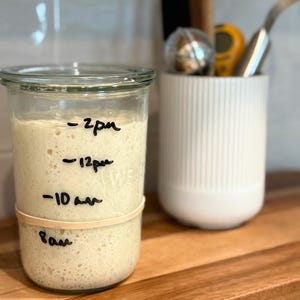 May include: A clear glass jar filled with beige sourdough starter, featuring black handwritten time markers for 8am, 10am, 12pm, and 2pm on the exterior. A tan rubber band is wrapped around the glass to track the rising level of the starter.