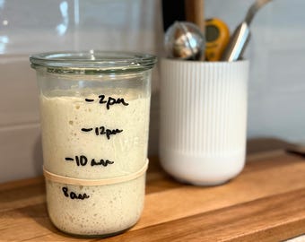 Puede incluir: Un frasco de vidrio transparente lleno de masa madre beige, con marcadores de tiempo negros escritos a mano para las 8am, 10am, 12pm y 2pm en el exterior. Una banda elástica color canela rodea el vidrio para seguir el nivel de la masa.