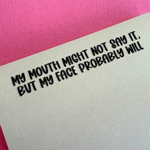 May include: A light beige card with the text "MY MOUTH MIGHT NOT SAY IT, BUT MY FACE PROBABLY WILL" in bold black letters. The card is set against a bright pink textured background.