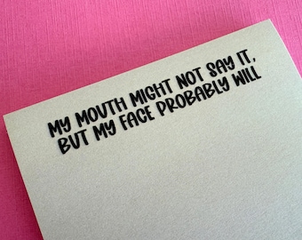 May include: A light gray rectangular card featuring the black printed text: MY MOUTH MIGHT NOT SAY IT, BUT MY FACE PROBABLY WILL. The card has a smooth, matte finish and is positioned against a bright pink textured background.
