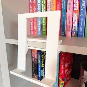 May include: A white, rectangular, open-frame book stand with a flat base and a central horizontal support bar. The stand is constructed from smooth, matte material and features a minimalist, upright design intended to hold a single book.