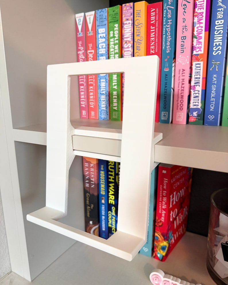 May include: A white, rectangular, open-frame book stand with a flat base and a central horizontal support bar. The stand is constructed from smooth, matte material and features a minimalist, upright design intended to hold a single book.