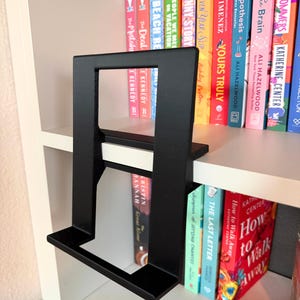 May include: A matte black, rectangular, L-shaped display stand featuring a wide base and a vertical support frame. The stand is positioned on a white shelf, with a collection of colorful books visible in the background behind the frame.