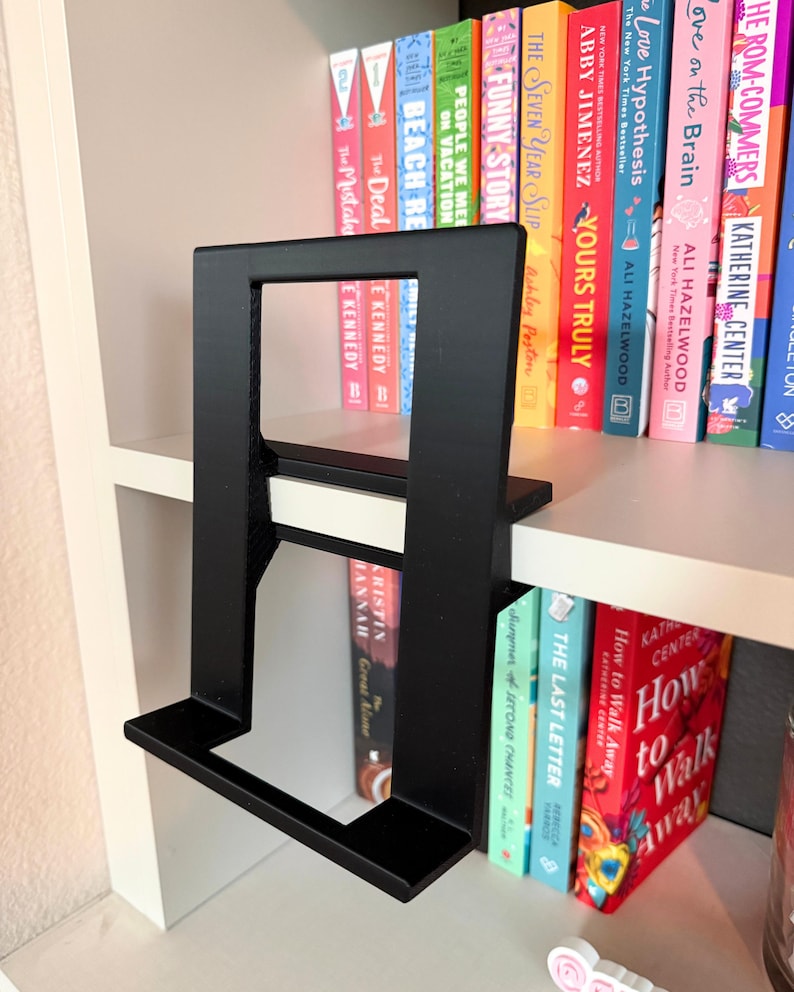 May include: A matte black, rectangular, L-shaped display stand featuring a wide base and a vertical support frame. The stand is positioned on a white shelf, with a collection of colorful books visible in the background behind the frame.