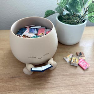 Op de afbeelding: Een matte beige, ronde houder met een glimlachend gezicht, voorzien van kleine armen die een open miniatuurboek vasthouden. De houder is gevuld met diverse kleine boekjes en er liggen nog enkele miniatuurboeken buiten de houder.
