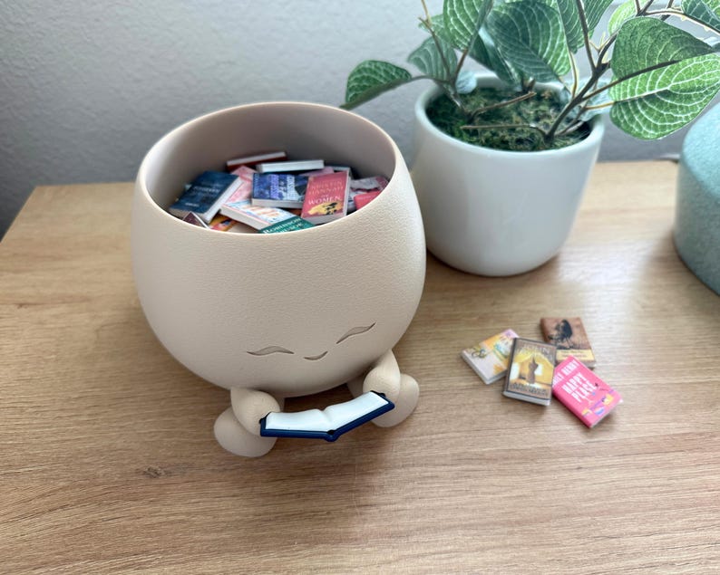 Op de afbeelding: Een matte beige, ronde houder met een glimlachend gezicht, voorzien van kleine armen die een open miniatuurboek vasthouden. De houder is gevuld met diverse kleine boekjes en er liggen nog enkele miniatuurboeken buiten de houder.