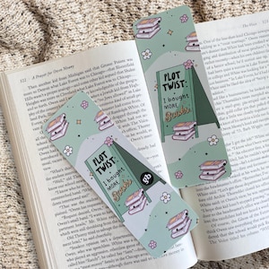 May include: Two green and white bookmark designs with a stack of books and the text "Plot Twist: I bought more books". The bookmarks have a floral pattern and are on a beige fabric background.