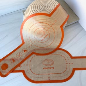 May include: Two orange silicone baking mats with circular and oval designs, and the text "KOOLSTUFFS". One mat is partially rolled, revealing a light brown, coiled object. The mats feature measurement guides in inches and centimeters.