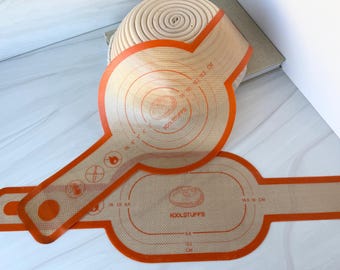 May include: Two beige silicone baking mats with orange borders and handles. The mats feature printed circular and oval measurement guides in inches and centimeters, along with the text KOOLSTUFFS and various icons indicating heat and usage guidelines.
