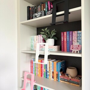 May include: A white bookshelf featuring three L-shaped book display stands in black, white, and pink. The shelves are filled with various hardcover and paperback books, a small potted green plant, and a decorative sign on a miniature easel.