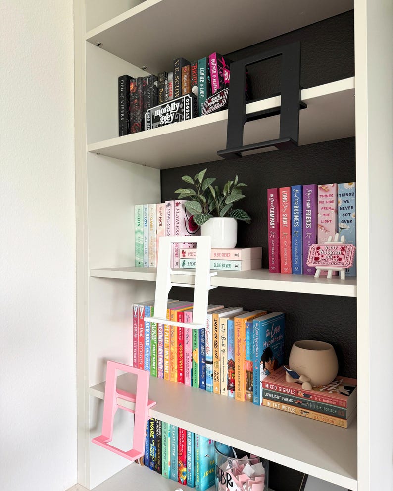 May include: A white bookshelf featuring three L-shaped book display stands in black, white, and pink. The shelves are filled with various hardcover and paperback books, a small potted green plant, and a decorative sign on a miniature easel.