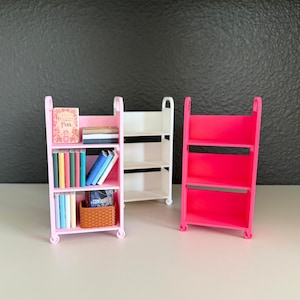 May include: Three miniature rolling utility carts with three shelves each. One light pink cart is filled with small books and a brown woven basket. One white cart is empty. One bright pink cart is empty. Each cart features four small wheels.