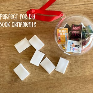 Puede incluir: Una bola transparente llena de r&eacute;plicas de libros en miniatura, con una cinta roja para colgar. Varios bloques rectangulares blancos est&aacute;n dispersos alrededor de la bola. El texto "PERFECT FOR DIY BOOK ORNAMENTS" es visible.