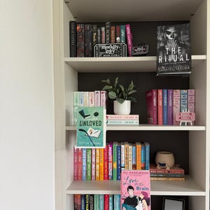 May include: A white bookshelf filled with numerous books arranged on five shelves. The collection includes various titles, a small potted plant, decorative book-themed signs, and two books displayed on small stands facing forward, titled Unloved and Love on the Brain.