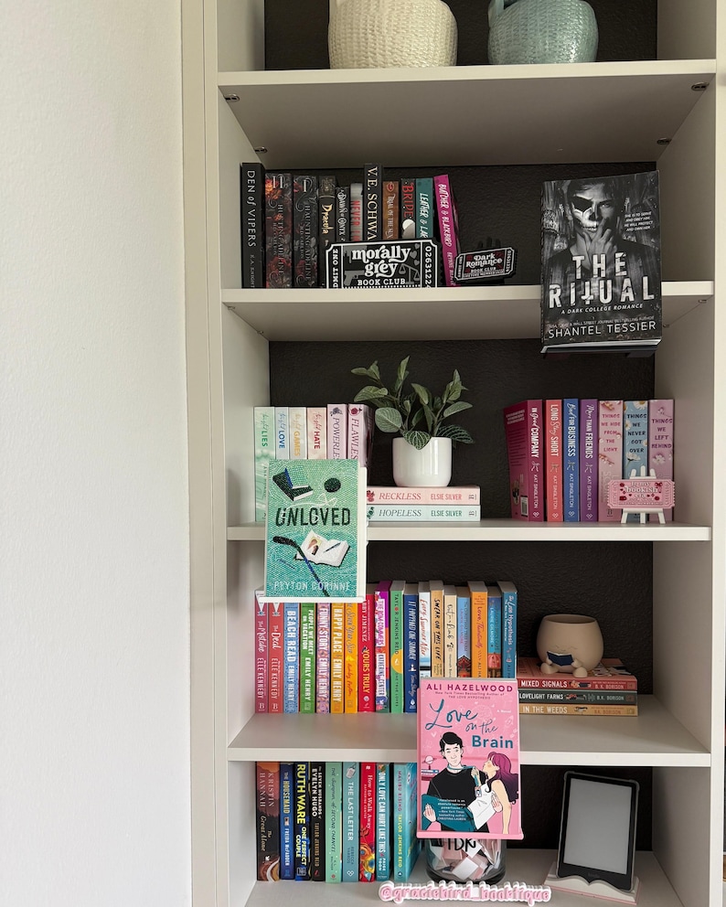May include: A white bookshelf filled with numerous books arranged on five shelves. The collection includes various titles, a small potted plant, decorative book-themed signs, and two books displayed on small stands facing forward, titled Unloved and Love on the Brain.