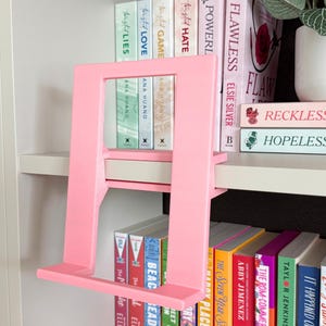 May include: A light pink, rectangular book stand with a flat base and an angled support frame. The stand is constructed from a solid material and features a minimalist, open-center design, positioned to hold a book upright on a shelf.