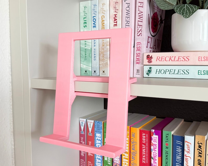 May include: A light pink, rectangular book stand with a flat base and an angled support frame. The stand is constructed from a solid material and features a minimalist, open-center design, positioned to hold a book upright on a shelf.
