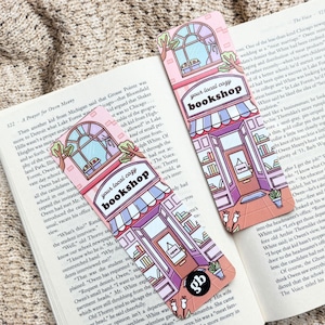 May include: Two pink and purple bookmarks with the text "your local cozy bookshop" are displayed on an open book. The bookmarks feature a bookstore illustration with books, a window, and an open door.