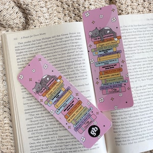 Peut inclure: Deux marque-pages rectangulaires roses illustrés d'une pile de livres colorés surmontée d'un chat gris endormi. Les livres portent des titres comme The Book Whispered, Pages and Peace et Cozy Chronicles. Des fleurs et étoiles blanches décorent le fond.