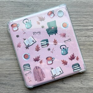 May include: A clear tablet case with a pink background featuring illustrations of autumn-themed items. The design includes a green armchair, books, eyeglasses, candles, teacups, a teapot, and fall leaves.