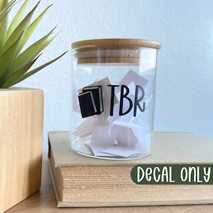 May include: A clear glass jar with a bamboo lid, filled with white paper scraps. The jar has a black decal with the letters "TBR" and a book icon. The jar sits on a vintage book. A wooden planter with a green plant is to the left.