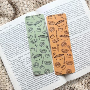 May include: Two rectangular bookmarks featuring a repeating black line art pattern of kitchenware, including Dutch ovens, bowls, and whisks. One bookmark is sage green, and the other is a warm orange, both resting on an open book page.
