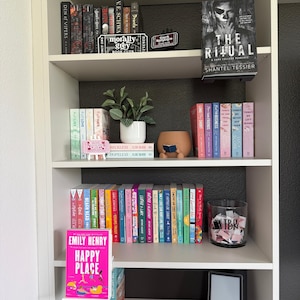May include: A white bookshelf filled with various books, a small potted plant, a brown ceramic figure reading a book, a glass jar labeled TBR containing paper slips, and a black e-reader. The book Happy Place by Emily Henry is displayed.