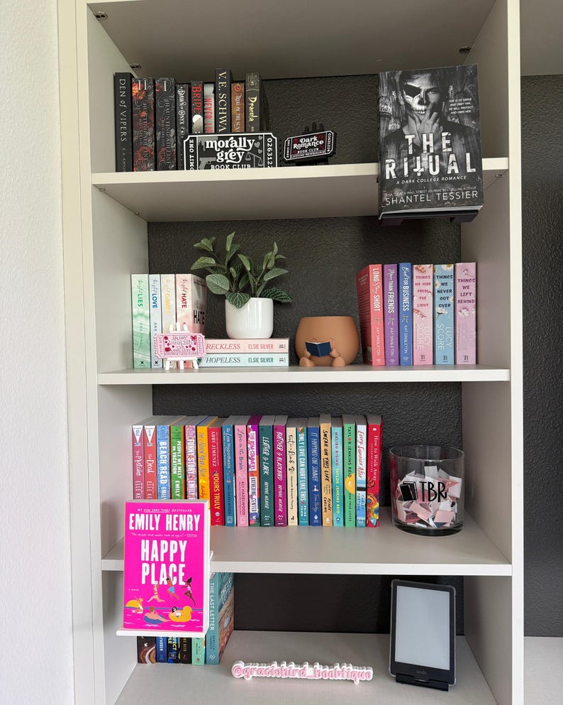 May include: A white bookshelf filled with various books, a small potted plant, a brown ceramic figure reading a book, a glass jar labeled TBR containing paper slips, and a black e-reader. The book Happy Place by Emily Henry is displayed.
