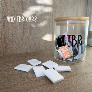 Puede incluir: Un tarro de cristal transparente con tapa de bamb&uacute;, etiquetado "TBR" con un gr&aacute;fico negro de libros, lleno de libros peque&ntilde;os. Objetos cuadrados blancos est&aacute;n esparcidos frente al tarro. El texto "AND TBR JARS" est&aacute; arriba.