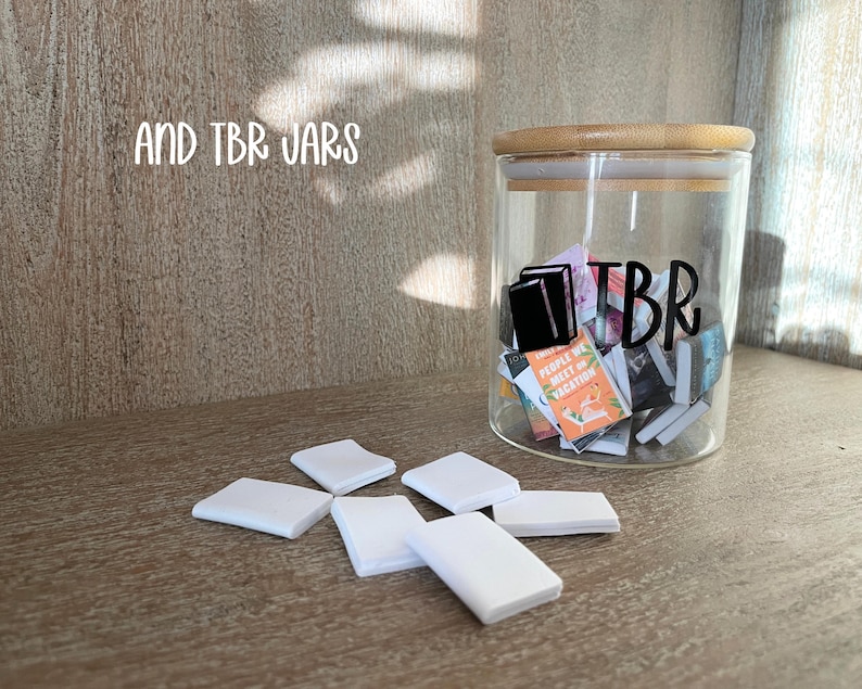 Puede incluir: Un tarro de cristal transparente con tapa de bamb&uacute;, etiquetado "TBR" con un gr&aacute;fico negro de libros, lleno de libros peque&ntilde;os. Objetos cuadrados blancos est&aacute;n esparcidos frente al tarro. El texto "AND TBR JARS" est&aacute; arriba.